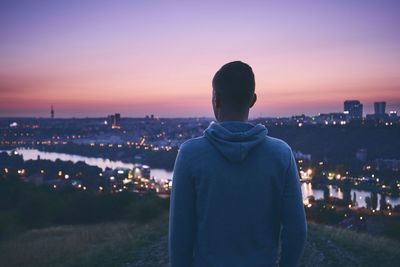 Rear view of man looking cityscape against sky at dusk