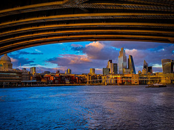 View of city buildings by river against cloudy sky