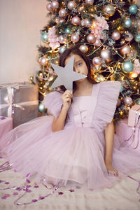 Beautiful baby girl with big eyes in a pink dress is sitting at the christmas tree at home hold star