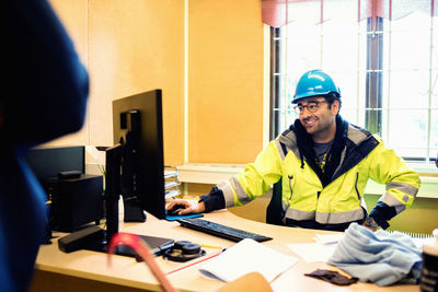 Smiling construction worker in protective wear talking to man while sitting at desk