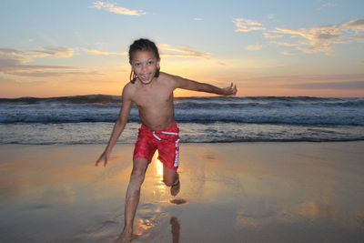 Portrait of smiling shirtless boy running on sea shore against sky during sunset