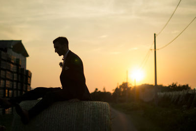 Side view of man standing by hay bale against orange sky