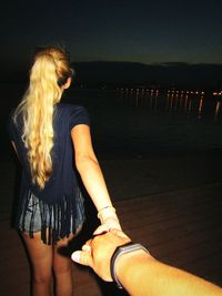 Cropped image of boyfriend holding hand of girlfriend while standing on shore at night