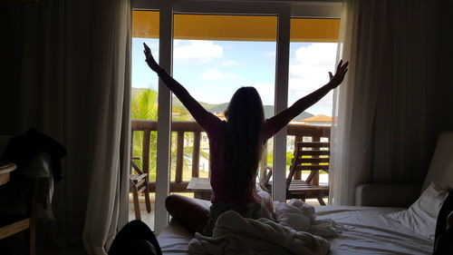 Rear view of woman with arms outstretched sitting on bed against window at home