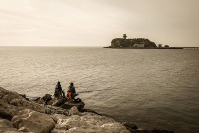 Rear view of people fishing in sea while sitting on rock against sky