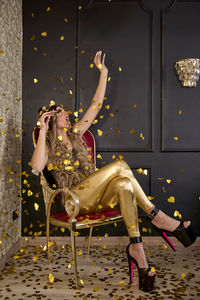 Non binary person playing with golden confetti sitting on chair at home