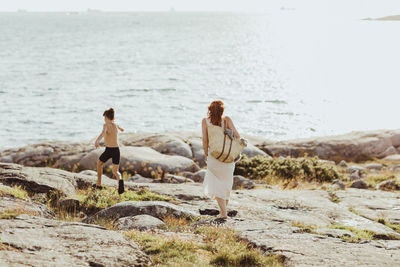 Rear view of woman with bag while son running against sea during sunny day