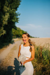 Portrait of happy bride sticking out tongue while standing with bouquet standing on country road