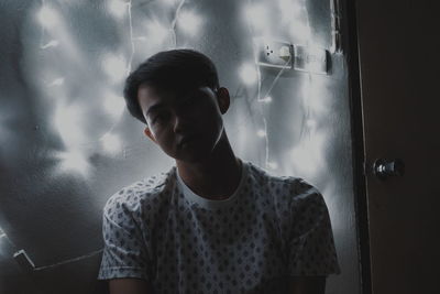 Portrait of young man standing by illuminated string lights on wall in darkroom