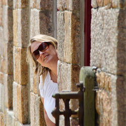 Portrait of woman wearing sunglasses while peeking through window on stone wall