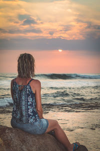 Woman sitting on rock at beach against sky during sunset