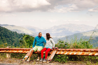 Couple sitting on fence against cloudy sky