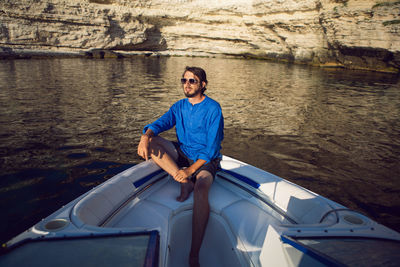 Man with a beard in blue shirt and sunglasses sitting on a yacht near the rocks in the sea in summer