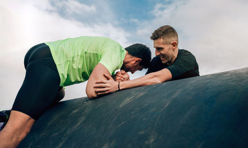 Man assisting friend in climbing on pipe against sky