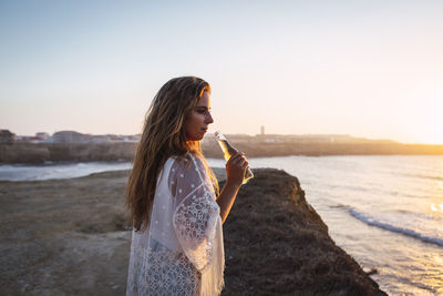 Young woman with small white wine bottle looking away while standing at beach
