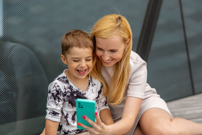 Happy blond woman and little boy sitting on terrace and making selfie on smartphone. mother and son