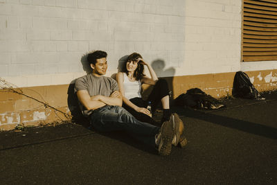 Male and female friends sitting side by side against wall on footpath