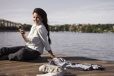 Portrait of happy businesswoman listening music while sitting on boardwalk at seaside