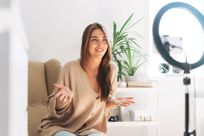 Young pretty woman blogger in cozy cardigan records video on mobile phone using ring lamp 