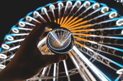 Cropped hand holding crystal ball against illuminated ferris wheel at night