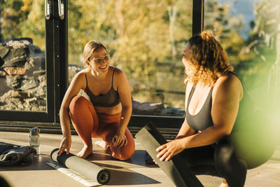 Happy female friends talking to each other while rolling exercise mats at retreat center