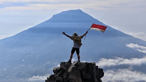 Rear view of man holding flag while standing on rock against sky