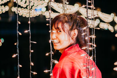 Portrait of smiling young woman with illuminated string lights at night