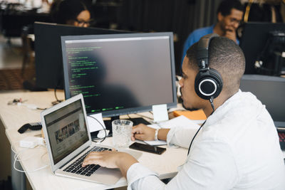 Side view of male programmer with headphones coding over laptop on desk while sitting in office