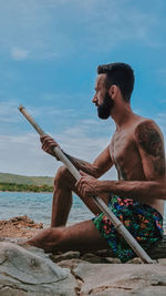 Full length of shirtless man sitting at beach against sky