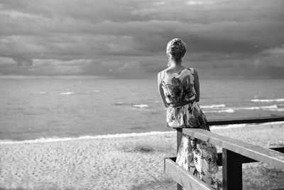 Rear view of woman sitting on railing at beach against cloudy sky