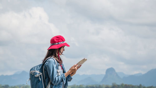 Side view of young woman reading map while standing against sky