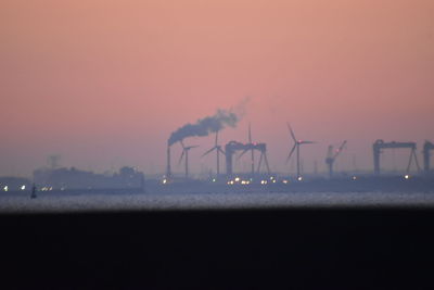 Silhouette factory by sea against orange sky