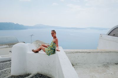 Young woman sitting on retaining wall by sea against sky