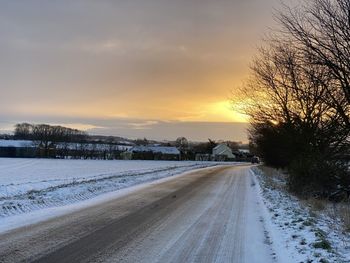 Road amidst snow covered plants against sky during sunset