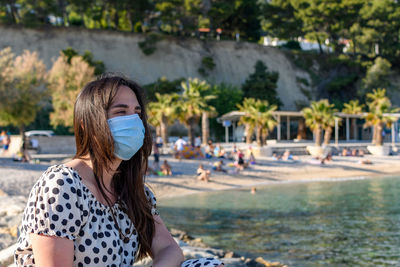 Young woman wearing surgical mask, sitting and relaxing on beach in summer.