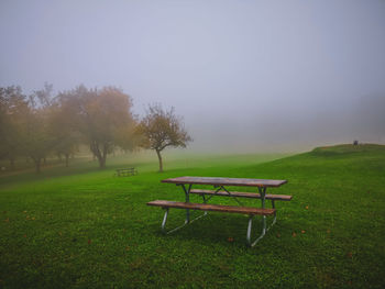 Empty bench on field by trees against sky