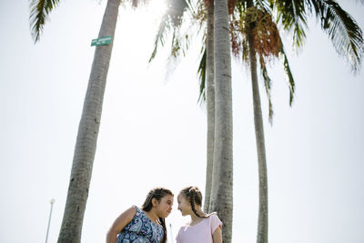 Sisters looking each other face to face while standing by coconut palm trees at beach against clear sky during sunny day