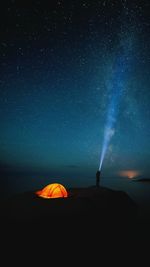 Silhouette person with flashlight standing by illuminated tent against star field at night