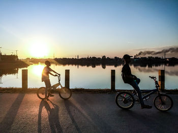 Boys riding bicycle at lakeshore against sky during sunset
