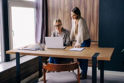 Two female colleagues are discussing a project in the office at the workplace by the window.