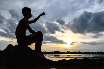 Silhouette boy sitting on rock against sky during sunset