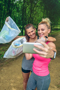 Female friends taking selfie while holding garbage in plastic bags at park