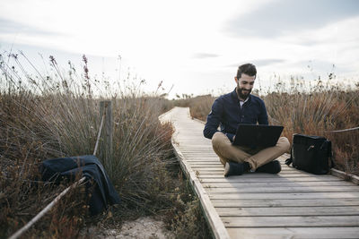 Full length of man using laptop while sitting amidst field on boardwalk