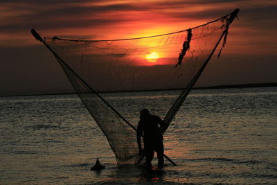 Silhouette fisherman with fishing net in sea against sky during sunset