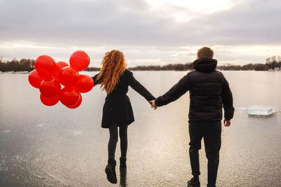 Rear view of couple holding balloon walking on frozen lake against sky