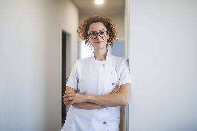 Positive young female therapist in white uniform and glasses smiling and looking at camera while standing in corridor of modern clinic