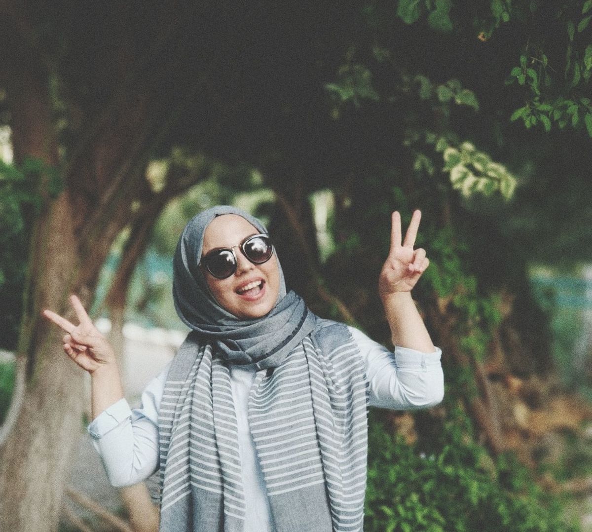 one person, front view, real people, gesturing, standing, portrait, lifestyles, glasses, waist up, leisure activity, sunglasses, focus on foreground, hand sign, casual clothing, emotion, symbols of peace, young adult, tree, peace sign, fashion, outdoors
