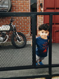 Portrait of cute boy standing by metallic fence against house