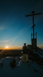 Young man in ski suit stands on mountain next to cross and looks at the sunset