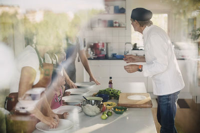 Senior man in chef's jacket guiding family in preparing asian food at kitchen